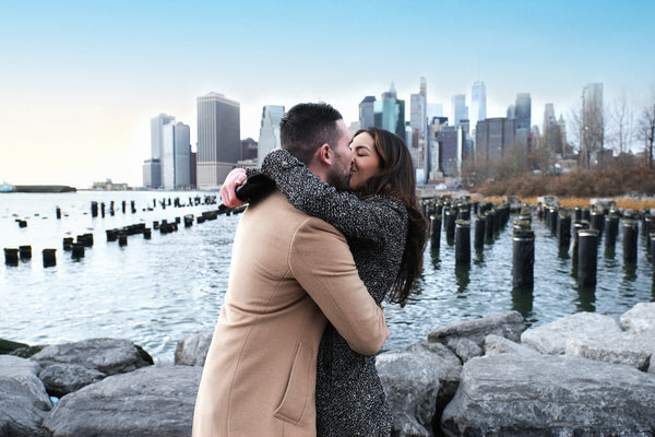 Engaged couple hugging on the Brooklyn waterfront rocks with the Manhattan skyline, NYC engagement photography