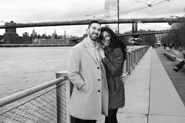 Black and white portrait of an engaged couple on a riverside promenade with the Brooklyn Bridge behind, NYC engagement photographer