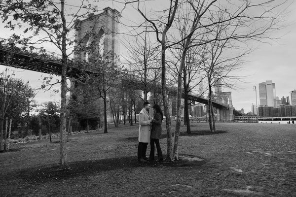 Black and white photo of a couple walking under bare trees near the Brooklyn Bridge, NYC engagement session by Lena Di