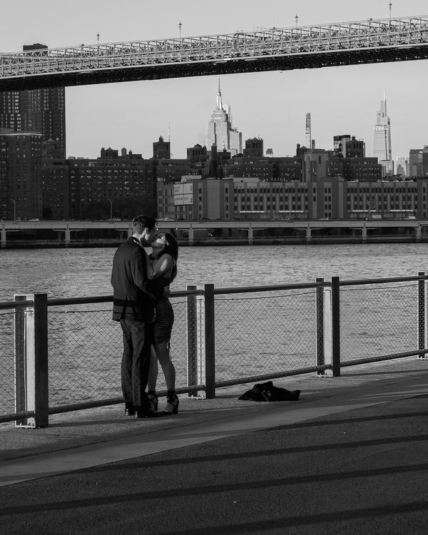 Black and white photo of a couple kissing at the Brooklyn waterfront railing under the Brooklyn Bridge with the Manhattan skyline, NYC engagement photographer Lena Di