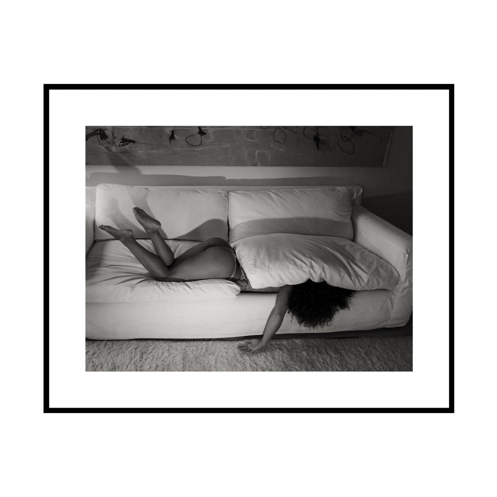 Hidden, black and white figure half-hidden under sofa cushions fine art photography print by Lena Di, shown framed with a white mat