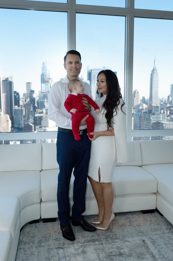 Family photographer NYC: father and mother standing in a white sofa penthouse with their baby in a red outfit, Manhattan skyline behind, by Lena Di