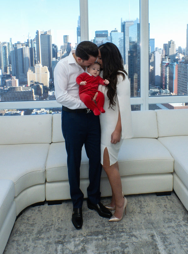 Family photography in New York City: father kissing his baby in a red outfit beside his wife by floor-to-ceiling skyline windows