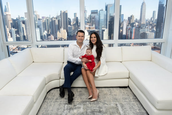 NYC family photographer: parents seated on a white sectional with their baby in a red outfit, city skyline windows, by Lena Di